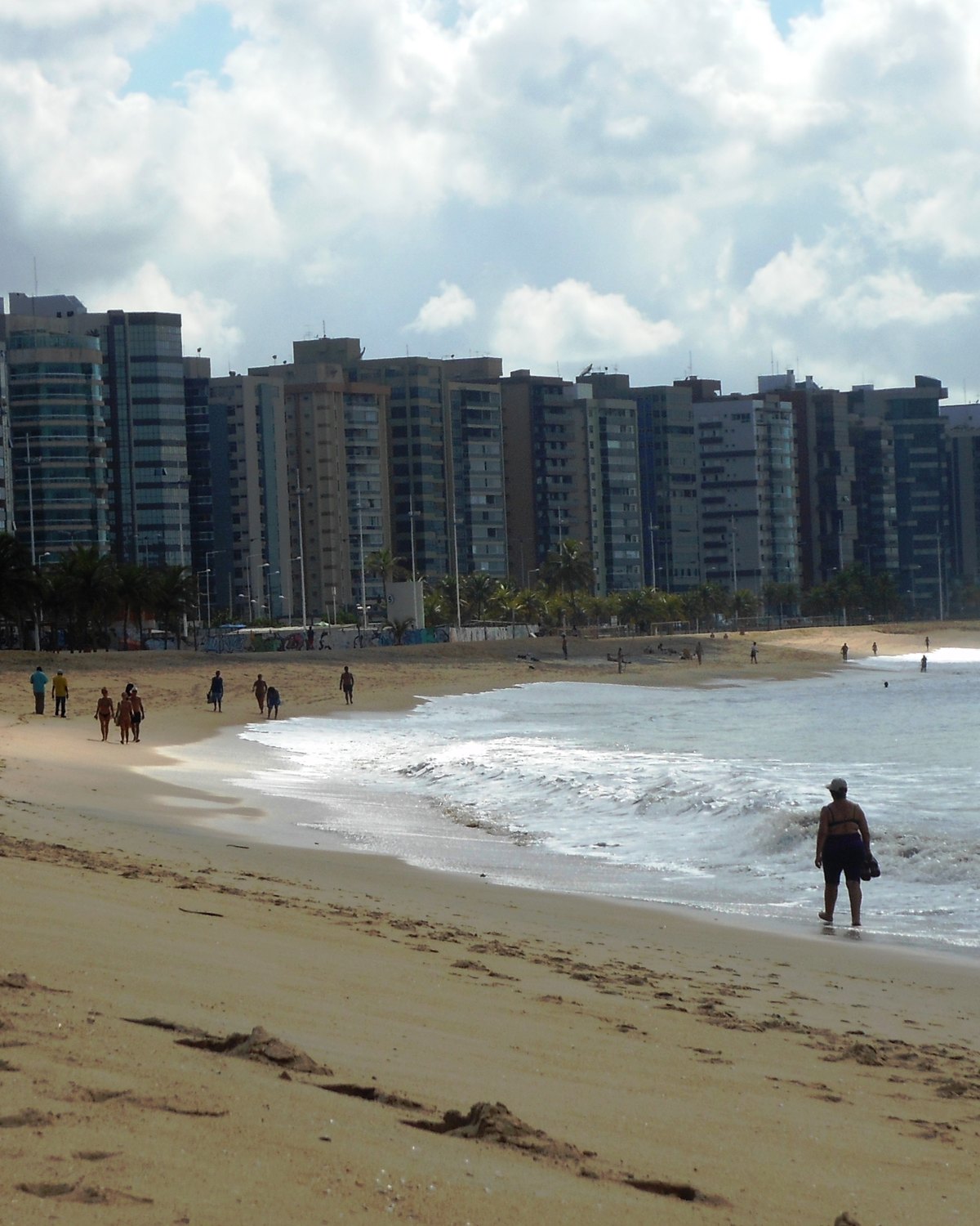 Pessoas caminhando na areia da Praia de Camburi, com a fileira de prédios da orla ao fundo.