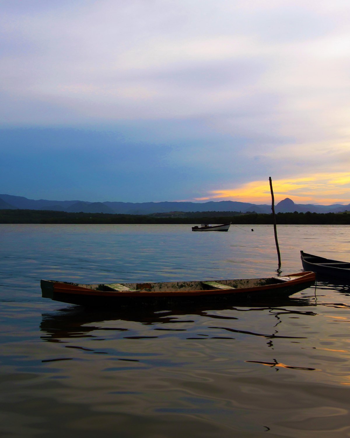 Canoas de madeira paradas na água da baía ao entardecer, na Ilha das Caieiras, com o morro recortado no horizonte.