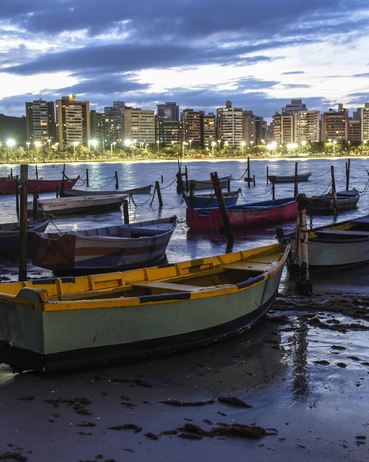 Barcos coloridos apoiados na maré baixa em frente à Praia do Canto, com as luzes dos prédios acesas no começo da noite.