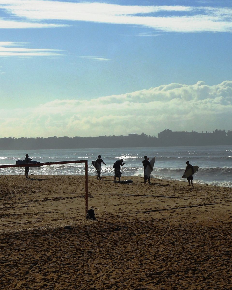 Surfistas atravessando a areia da Praia de Camburi com as pranchas debaixo do braço, em contraluz no fim da tarde.