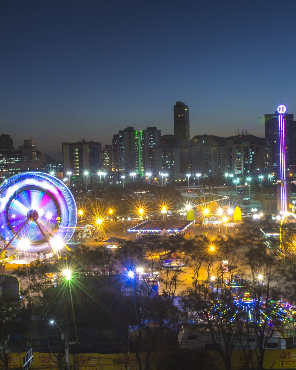 Parque de diversões iluminado à noite na Enseada do Suá, com roda-gigante e brinquedos acesos e os prédios de Vitória ao fundo.