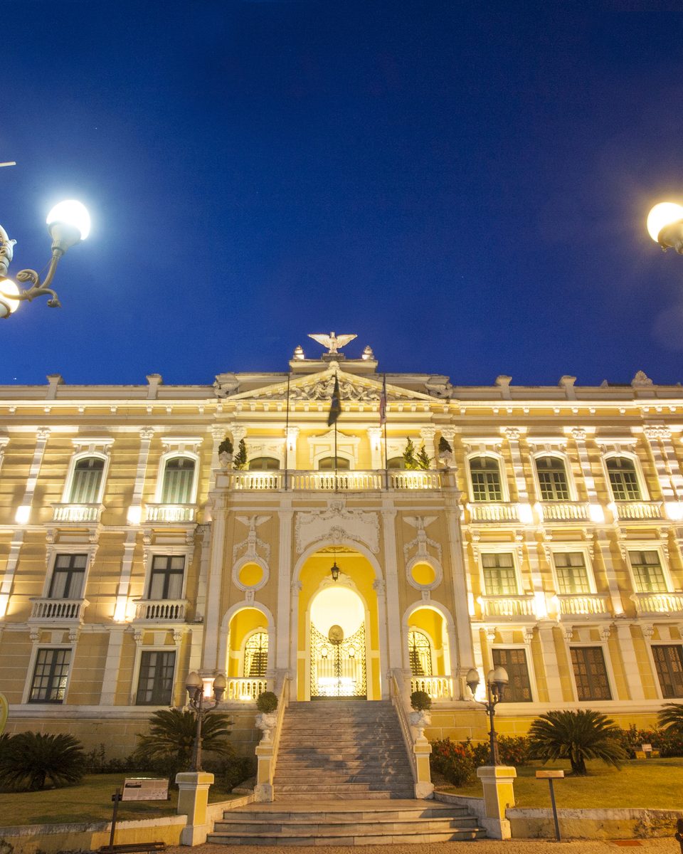 Palácio Anchieta iluminado ao anoitecer, com a fachada amarela e branca, a escadaria de acesso e dois lampiões antigos em primeiro plano.