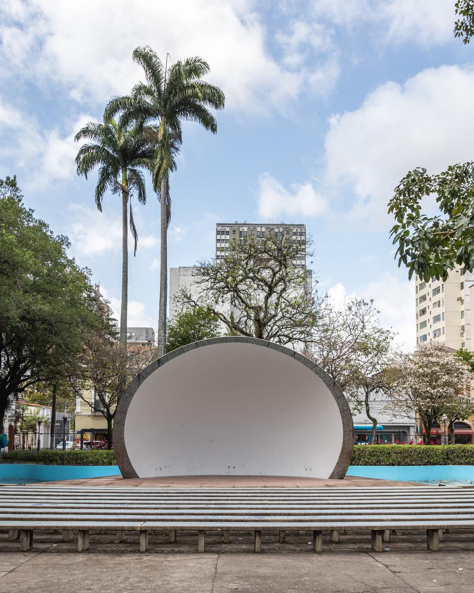 Concha Acústica branca do Parque Moscoso, com bancos de concreto em frente, palmeiras altas atrás e pessoas sentadas à sombra.