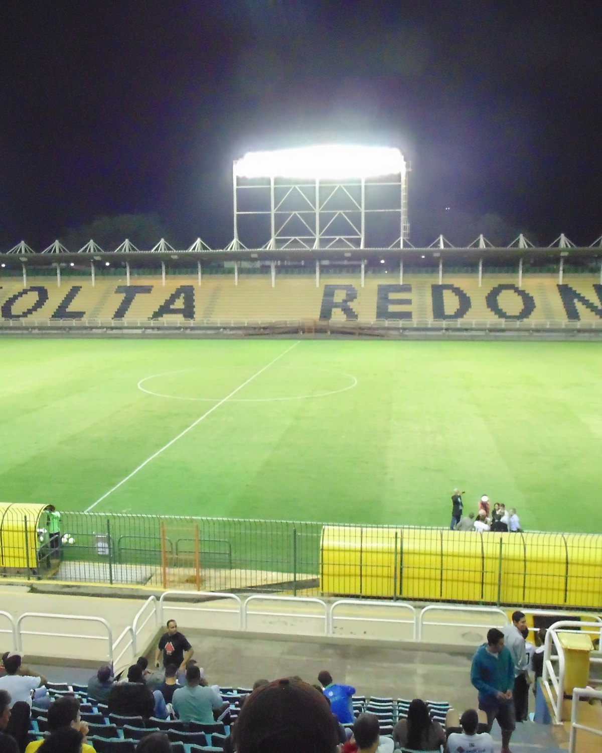 Estádio Raulino de Oliveira à noite, com os refletores acesos, o nome Volta Redonda escrito nas cadeiras amarelas da arquibancada do outro lado do gramado e torcedores sentados em primeiro plano.