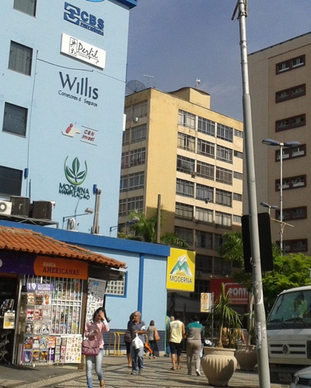 Calçada movimentada na Vila Santa Cecília, em Volta Redonda, com banca de revistas e pessoas passando.