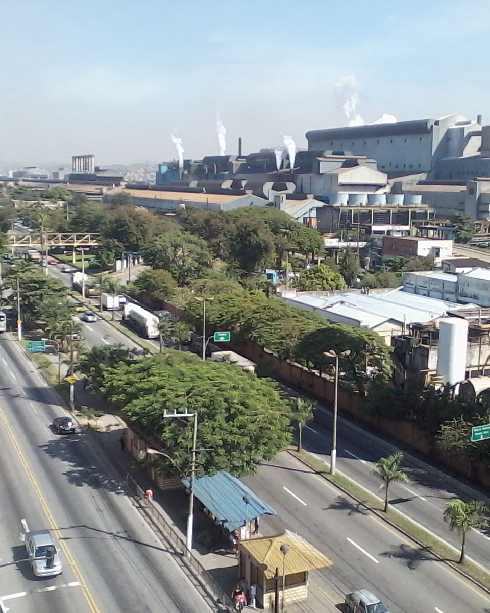 Avenida larga no centro de Volta Redonda vista do alto, com ônibus e carros, árvores no canteiro, um ponto de ônibus e os galpões e chaminés da CSN ao fundo num dia de céu azul.